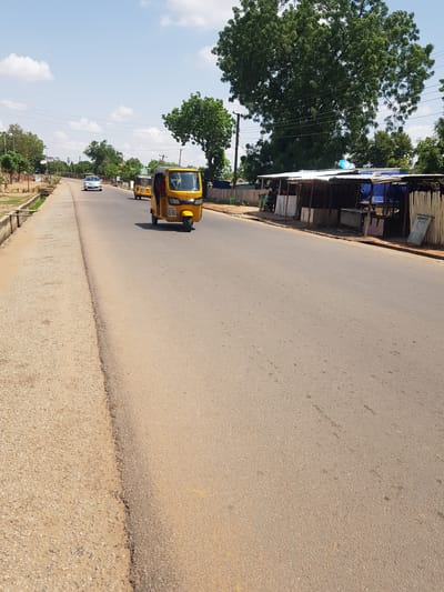 This media file depicts a roadside scene in Palade, Nigeria. The image is oriented vertically, with the road occupying the majority of the frame. The road surface appears to be asphalt, with a rougher, gravelly shoulder on the right side.

Several vehicles are visible. A bright yellow auto-rickshaw, also known as a "Keke Napep" or "Maruwa," is the most prominent vehicle, seen from the side and slightly from the front. It has a visible license plate with some text. Further down the road, another similar auto-rickshaw is visible, and a white sedan is also on the road.

On the left side of the road, there are structures that appear to be market stalls or small shops. They are constructed with corrugated metal roofs and wooden or metal framing, some covered with blue tarps. A large, leafy tree with dense foliage is prominent on the far left, with power lines running through its branches. More trees and vegetation are visible along the roadside and in the distance.

The sky is a bright blue with scattered white clouds, suggesting a clear and sunny day, likely in the late morning or afternoon. The overall impression is of a typical day on a road in a town or city in Nigeria, with everyday transportation and local commerce taking place. No specific events or emotional cues are evident beyond the normalcy of the scene.