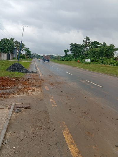 The image shows a road in Igrita, Nigeria under overcast skies. Road construction or repair is evident on the left side, with piles of dark gravel and disturbed reddish-brown earth. A broken concrete barrier and exposed rebar suggest ongoing work. A light pole stands near the construction zone. The road surface is wet and marked with worn yellow lines. In the distance, a truck and a motorcyclist are traveling on the road. A person wearing an orange shirt is walking away from the camera on the right side of the road, near a sign placed next to trees and green vegetation.