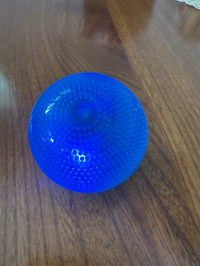 The image shows a close-up, overhead view of a textured, bright blue ball resting on a wooden surface. The ball is round and appears to be made of a translucent material, possibly plastic or rubber, with a dimpled surface that creates a pattern of small circular indentations. Lights are visible within the ball, casting a bright glow and highlighting the textured surface. The wooden surface beneath the ball has a natural grain pattern, suggesting it is a table or countertop. The lighting in the image is artificial, likely from the lights within the ball and possibly ambient room lighting. There are no people visible in the image. The scene is static, with no specific activity taking place. The overall impression is a simple, focused shot of an object. The provided location context "Sant'Angelo di Piove di Sacco, Italy" does not offer any visual cues present in the image itself. There is no visible text in the image.