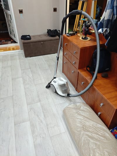 A wide shot captures an indoor scene, likely a home entryway or hallway. The floor is covered with light-colored laminate flooring with a wood grain pattern. A white and gray canister vacuum cleaner with a silver extension wand and a black hose is positioned on the floor. The hose snakes towards a wooden furniture piece that has multiple drawers and a bench-like surface. On the bench, a dark blue backpack with the word "VIVO" is visible. Beside the backpack, a small, dark brown spherical object rests. To the left of the vacuum, a brown storage bench with a tufted lid is present. A black folded garment sits on top of the bench. A closed door with a white patterned grate is visible to the far left of the frame. The wall is painted a neutral, light color. A small, square light fixture is mounted on the wall above the storage bench. Further to the right, a wooden cabinet with drawers is visible. On top of the cabinet, a decorative item featuring a black figurine holding a gold plate is displayed, along with a toy car and some other small objects. A folded blue and white plaid garment hangs from a rod to the right of the cabinet. In the foreground, a rolled-up carpet, wrapped in clear plastic, lies on the floor. The scene does not contain any indications or elements that specifically place it within the city of Arshaly, Kazakhstan.