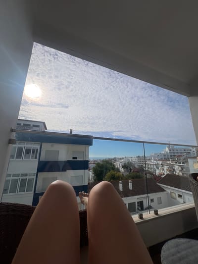 A close-up, low-angle shot shows a person's legs in the foreground, positioned on a wicker chair on a balcony. The legs are bare, and one foot is visible, painted with light blue nail polish. The backdrop is an urban landscape of white buildings with red-tiled roofs, characteristic of Lagos, Portugal. The sky above is a bright blue, filled with wispy white clouds, suggesting a sunny day. The sun is shining, creating lens flare in the bottom left corner of the image. The scene is captured from a balcony, indicated by the presence of a glass railing and the viewpoint looking out over buildings. The overall mood is relaxed and summery, implying a leisurely moment on vacation. There is no visible text in the image.