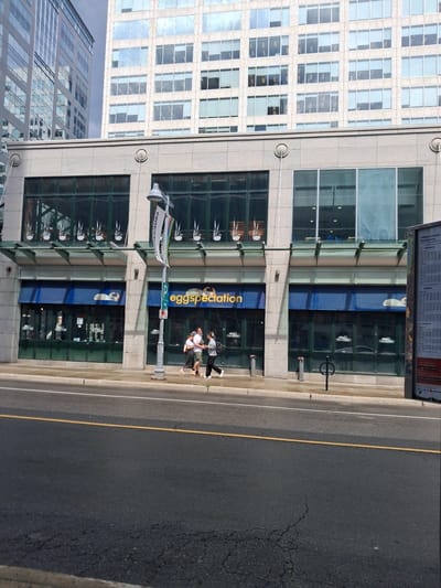 The image shows a street view in Ottawa, Canada, featuring a modern building with large glass windows. Two people are walking on the sidewalk in front of the building. The building has a blue awning with the text "eggspectation" visible. Above the awning, there are more windows, some displaying white graphics of what appear to be birds. A street lamp with a sign that reads "centretown" is also visible. The street is paved with asphalt and marked with yellow and white lines. The sky appears bright, suggesting it is daytime. The reflection in the building's windows indicates a clear, sunny day. The overall scene conveys a sense of urban activity and a bright, pleasant atmosphere.