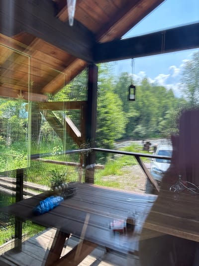 The image captures a view from inside a wooden structure, likely a porch or balcony, looking out towards a natural landscape. The foreground features a wooden table with a blue plastic-wrapped object and a planter with small potted plants. Reflections on the glass surface obscure some details of the interior.

Outside, a verdant forest surrounds a body of water, possibly a river or stream, with a white car parked nearby. The sky is partly cloudy with a bright blue hue. A rustic wooden railing separates the porch from the outdoor area. The architecture of the structure suggests a rustic, possibly remote, setting. The presence of a car indicates accessibility to the location. The overall scene is one of a tranquil, nature-adjacent environment, characteristic of areas outside of major urban centers.