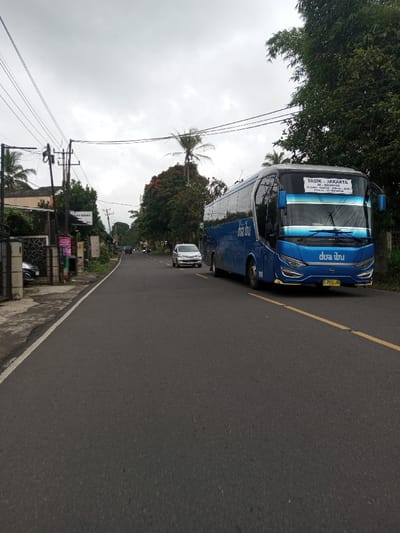 The scene depicts an asphalt road in the city of Kodarapalli, India, under an overcast sky. A large, blue passenger bus, branded "dua ibu" on its side and front, occupies the right side of the road, facing the viewer. The bus displays "TASIK - JAKARTA" as its destination on the front sign, with additional smaller text below, and bears an Indonesian license plate "Z 7536 AW". Behind the bus, a silver minivan follows on the same side of the road, and other vehicles are visible in the distance. The road features a yellow solid line dividing the visible lane and a white line marking the left edge. To the left of the road, a narrow paved shoulder or sidewalk borders a dark metal fence. Beyond the fence, residential or commercial buildings are visible, partially obscured by trees and foliage. One building has a sign with "SAAT SEHAT" text. Multiple utility poles with overhead lines run along the left side. The right side of the road is lined with dense green trees and vegetation, including visible palm fronds. No individuals are clearly discernible.