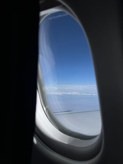 The media file captures a view from an airplane window, showcasing the vast blue sky and distant clouds from high altitude. The image is composed with the dark interior of the aircraft framing the oval-shaped window, which acts as the main viewport.

Through the window, a bright, clear blue sky dominates the upper and left portions of the visible exterior. Along the right side, an airplane wing is partially visible, indicating the aircraft's movement. Below the wing and stretching across the horizon, a layer of thin, wispy white clouds can be seen. The clouds appear somewhat scattered or in lines, suggesting a relatively clear sky above them.

The bright sunlight illuminating the sky and clouds indicates that the image was taken during daytime. There are no people visible in the frame, and the focus is entirely on the aerial landscape. Faint, blurry text, possibly a label or instruction, is barely discernible on the window's surface, but it is illegible. The setting implies active flight over a geographic area, with the provided context being Tsagaanchuluut, Mongolia. The scene evokes a sense of travel and the expansive beauty of the sky from above.