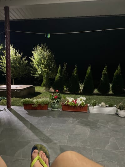 The image depicts a night-time scene in the courtyard of a residential property in Slobozia-Dușca, Moldova. The foreground shows a person's legs and feet, clad in a blue and green flip-flop, resting on a tiled patio. The patio is illuminated by artificial light, casting long shadows from a nearby pillar and roof. Beyond the patio, a well-maintained lawn is visible, lined with various potted plants and small evergreen trees. In the background, a clothesline with two clothespins is strung across the darkness. The overall ambiance suggests a quiet, outdoor evening.