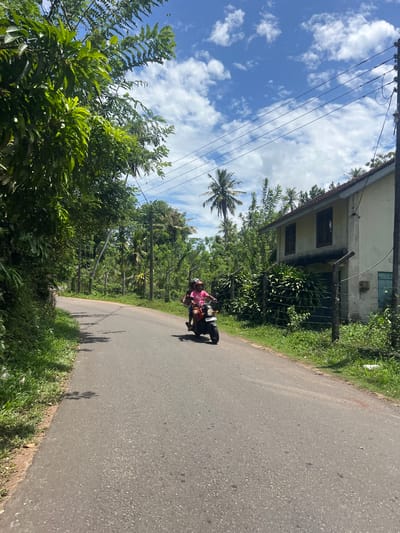 The image depicts a road in Moodugamuawa, Sri Lanka, flanked by dense vegetation on both sides. A single-story building is visible on the left side of the road amidst the foliage. Overhead electrical wires are strung across the sky, which features a mix of blue and white clouds. A motorcycle with two riders is traveling along the road. The riders are wearing casual clothing, with the front passenger wearing pink. The road surface is asphalt. The scene is captured in daylight.