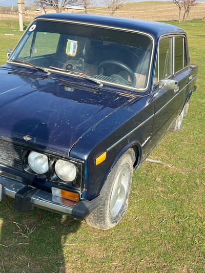 A dark blue, four-door sedan, likely a Lada (VAZ) model, is parked stationary on a grassy field in Qoraobdol QFY, Uzbekistan. The vehicle is angled, showing its front and driver's side. The paint is faded and covered in dust and dirt, with visible scratches and imperfections on the hood and fenders. The front bumper is chrome with black rubber end caps. Two round headlights and amber square turn signals are visible. A small Uzbek flag and what appears to be a rosary hang from the rearview mirror inside the dirty windshield. A white sticker with a black number "4" is affixed to the upper left corner of the windshield. The interior is partially visible, showing a dark steering wheel, possibly with a textured cover. The driver's side front wheel has a silver-colored, dirty hubcap or alloy wheel and a worn tire. The ground is a mix of green grass and patches of dry earth, suggesting an uncultivated area. In the background, scattered deciduous trees or bushes are visible, devoid of leaves, against a gently sloping, bare hill under a clear sky. A utility pole is present to the far left. No individuals or active interactions are depicted.