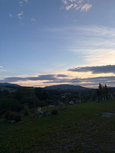 This image depicts a panoramic view of the outskirts of Limanowa, Poland, at dusk. The sky is a gradient of blues, transitioning to a warm orange and pink hue near the horizon, indicative of a sunset. Wispy clouds streak across the sky, some tinged with the sunset colors. Below the sky, rolling hills covered in dense, dark green trees form a prominent landscape. Scattered throughout the treeline and on the slopes are several houses with dark roofs, suggesting a residential area nestled within the natural environment. The foreground is dominated by a grassy slope with some scattered gravel. In the middle ground, closer to the houses, there are more trees and a few visible structures, including what appear to be garden elements and possibly outdoor recreational equipment. The overall impression is a serene, rural twilight scene.