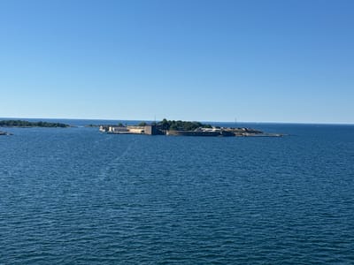 The image shows a clear blue sky and a calm blue sea. In the middle ground, there is a small island or peninsula with several buildings and fortifications. The structures appear to be made of stone and have a historical appearance, possibly part of a naval base or castle. Trees and vegetation are visible on the island. The water is rippled, suggesting a gentle breeze. The scene is devoid of people and other living beings. The location is the island of Tjärö, part of the Karlskrona archipelago in Sweden, famous for its naval heritage and fortifications.
