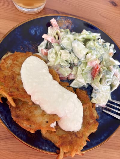 This media file shows a close-up, overhead shot of a plate of food on a wooden surface. The plate is dark blue with a gold rim and appears to be on a table.

The food consists of two main parts: a salad and potato pancakes topped with a dollop of sour cream or yogurt. The salad is composed of chopped lettuce, cucumber, tomatoes, and green onions, all mixed with a creamy dressing. The potato pancakes are golden brown and appear to be crispy. The sour cream is white and smooth, covering a significant portion of the pancakes. A fork is visible in the upper right corner, resting on the plate. To the left of the plate, a glass with a light amber liquid, possibly juice or a beverage, is partially visible.

The lighting suggests it is daytime, and there are no indicators of the weather. The setting appears to be a casual dining environment, possibly a home or a restaurant. There is no text visible in the image. The overall impression is of a wholesome and appealing meal.