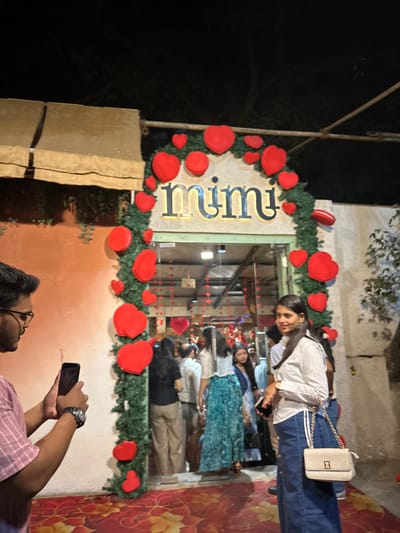 The scene depicts a nighttime outdoor view of a decorated establishment, identified by the illuminated sign "mimi" above its entrance. The entrance forms an archway heavily adorned with green foliage garlands and numerous red heart-shaped decorations of various sizes. The ground in the immediate foreground is covered by a red carpet with an orange floral pattern.
To the left, a male individual, seen from the right profile, wears glasses and a striped light-colored shirt. He holds a black smartphone horizontally, seemingly capturing a photograph or video of the decorated entrance. A wristwatch is visible on his left wrist.
To the right, a female individual stands, facing slightly towards the left but looking forward. She has dark hair and wears a light-colored, long-sleeved collared shirt and a dark denim skirt or dress. She carries a small, light-colored shoulder bag with a gold chain strap across her body and holds a smartphone in her left hand.
Inside and around the entrance, a crowd of approximately 10-15 individuals, mostly women, are visible, either entering, exiting, or congregating. One woman inside is notable for wearing a long teal-patterned skirt. The interior of the venue also features hanging red heart decorations, visible through the open doorway. To the far left, an orange-toned wall with subtle green decorative elements and an awning-like structure is visible. This scene is located in the city of Kalyan-Dombivli, India.