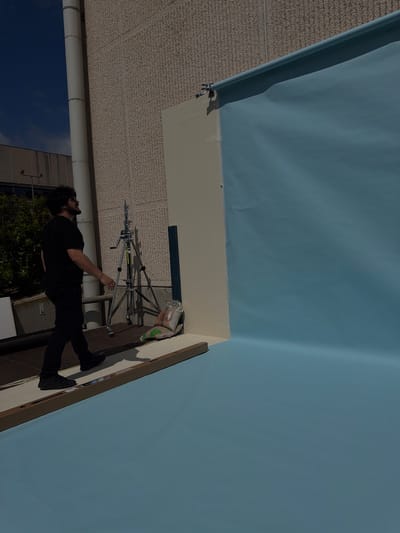 The image captures a man setting up a backdrop, possibly for photography or videography, outdoors. The backdrop is a large sheet of light blue material, partially draped and attached to a wall or building with what appears to be clips.
The man, dressed entirely in black, is positioned near the center of the frame, seemingly adjusting the equipment. A silver photography light stand is behind him.
The location appears to be an urban area, with a glimpse of a building with windows and greenery visible in the background. The scene is set against the backdrop of the sky. The exact location in Fibracolor, Spain, cannot be determined from this image alone.
