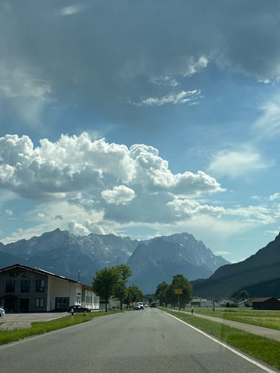 The image depicts a scenic view of a road leading towards a mountain range in Farchant, Germany. The sky is a mix of blue and grey clouds, with large, fluffy white clouds dominating the upper portion of the frame. The mountains are rugged and majestic, with some patches of snow visible on their peaks. In the foreground, a paved road stretches into the distance, bordered by green grass.
On the left side of the road, there is a building with "KFZ" signage, likely an automotive service center. Several trees are planted along the roadside, adding to the natural beauty of the scene. A few cars are visible on the road, moving away from the viewer. On the right side of the road, there are houses and fields, indicative of a rural or semi-rural setting. A yellow sign with the text "Farchant" is visible on the right side of the road, confirming the location. The overall impression is one of a peaceful, picturesque landscape, characteristic of the Bavarian Alps region.