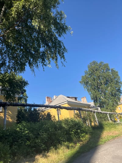 A bright, sunny day in Kiselyovsk, Russia, is depicted in this image. The scene features a yellow building with a grey roof and brick chimneys. A thick, insulated pipe runs horizontally across the mid-ground, supported by metal poles. Lush green trees and dense bushes surround the building, with a pathway visible on the right side of the frame. The sky is a clear, vibrant blue.