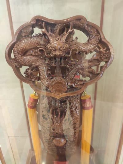The image displays a wooden carving of a dragon, intricately detailed and polished to a sheen. The dragon is positioned centrally, its head facing forward with ornate facial features and flowing mane. Its body curves around, suggesting movement. Below the dragon's head, a decorative element resembling a cluster of beads or spheres is carved. Hanging from the sides of this central piece are two yellow tassels, each adorned with a red and gold band near the top. The carving appears to be displayed within a glass case, as indicated by reflections and the overall clarity of the object. The background is out of focus, showing what appears to be a light-colored wall with vertical lines, possibly part of an interior display. There are no people visible in the image, and no specific location in Ürümqi, China, can be identified from the visual information provided.