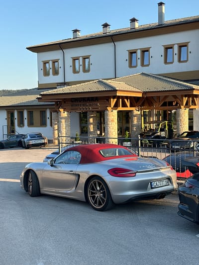 The image shows a view of the front entrance of the CHUKARA WINE & SPA HOTEL, located in Kerenite, Bulgaria. Several cars are parked in front of the hotel. In the foreground, a silver convertible Porsche Boxster with a red soft top is parked. Behind it, to the left, is a silver Porsche 911 with a Bulgarian license plate that reads "CB 6687 XN". Further left, a dark gray sports car is visible. Inside the hotel entrance, a black and neon green off-road vehicle is parked. The hotel has a white facade with wood accents and a tiled roof. The scene is illuminated by natural sunlight, indicating it is daytime.