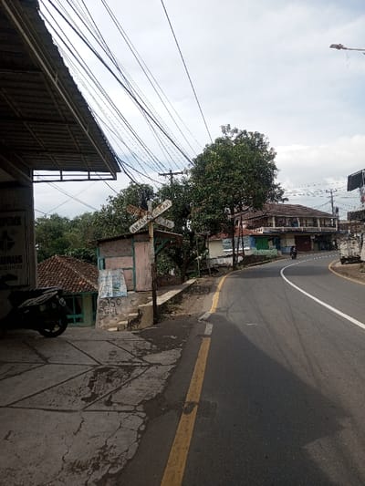 The scene depicts a roadside in the city of Chhenangri_Rishor, Bhutan, under an overcast sky. In the foreground, a dark-colored scooter is parked on a cracked concrete pavement beneath a metal-framed roof overhang extending from a building, a partially visible white sign on which reads "PT.IME" and "URUS". To the right of the scooter, a small, weathered wooden booth or shack, light-colored and featuring a corrugated roof, stands with posters affixed to its exterior. Adjacent to the shack, a white X-shaped sign mounted on a wooden post displays "AWAS" on the top plank and "KERETA API" on the bottom, indicating a warning.
An asphalt road with a solid yellow line on its right edge and a solid white center line curves to the right, leading into the background. A single individual on a dark-colored motorcycle is visible traveling on the road in the midground, heading around the curve. Along the right side of the road, multiple buildings are present, including a prominent two-story structure with a reddish-brown tiled roof and a balcony. Dense green trees and foliage are scattered throughout the image, both behind the structures on the left and along the curving road. Numerous utility poles and a complex network of overhead wires crisscross the sky. A white truck or utility vehicle is partially visible on the far right in the background.