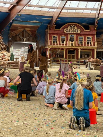 This image depicts an outdoor scene within a Western-themed attraction, likely part of a larger entertainment venue in Collendoorn, Netherlands. In the foreground, a group of children, predominantly girls, are seated on the ground, facing a wooden stage. They are wearing colorful feather headbands, suggesting participation in a themed activity. Some children are holding drumsticks and appear to be engaged in a drumming session with colorful plastic buckets arranged in front of them.

Behind the children, two adults dressed as Native Americans stand on the stage, facing the children. The adult on the left wears a brown buckskin outfit with a headdress and holds a feather. The adult on the right also wears a similar buckskin outfit and holds a drumstick, poised as if about to strike a drum. The stage area is decorated with elements reminiscent of the Wild West, including wooden structures, a wagon wheel, and a skull above an entrance.

Further in the background, a large, two-story building with a sign reading "BOOTHILL SALOON" is visible, further reinforcing the Western theme. A horse is tethered near the saloon, attended by a person in Western attire. The ground is covered in a light-colored sandy soil, littered with colorful confetti. The overall impression is that of a performance or educational activity designed for children, set within an immersive themed environment. The lighting suggests it is daytime, with natural light filtering through a partially covered roof structure made of wood and glass.