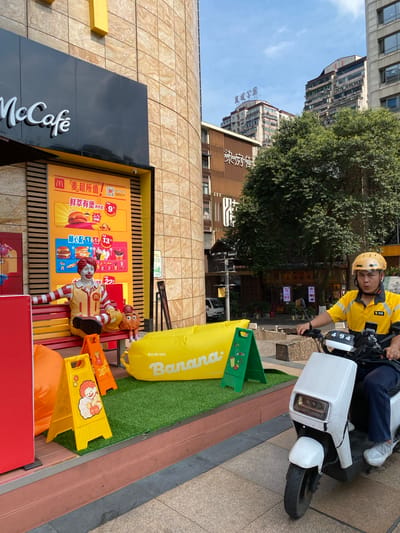 This is a medium shot, outdoor, eye-level photograph taken in the city of Jinjiang District, China. In the foreground, a person wearing a yellow uniform and a yellow helmet is riding a white electric scooter. The person is looking at the camera. In the midground, a McDonald's McCafe storefront is visible. A statue of Ronald McDonald is sitting on a red bench, next to a small statue of the Hamburglar. A large yellow inflatable banana is on the ground in front of the McDonald's. In the background, several tall buildings and trees are visible under a clear blue sky. The scene is illuminated by bright daylight.