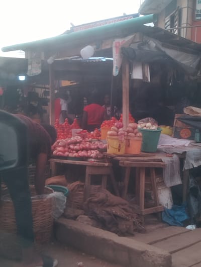 This is an image of a market stall in Aketu-Oja, Nigeria. The stall is covered by a green awning and is filled with various produce, including a large pile of red tomatoes and several buckets of onions. A person in a dark red shirt is bending over a basket to the left of the frame. Other people are visible in the background, partially obscured by the stall's structure. The market appears to be outdoors, with a rough, uneven ground surface. The overall atmosphere suggests a busy, local marketplace.