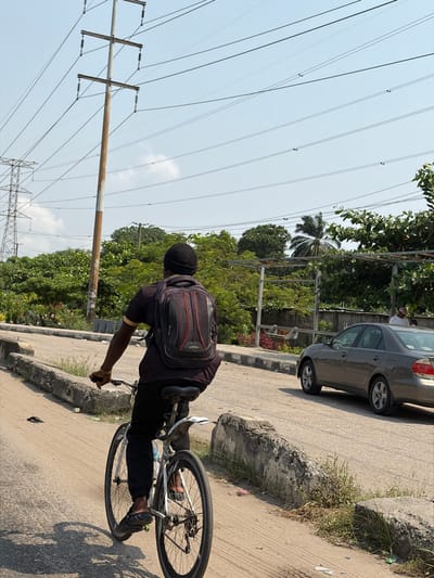 A male individual is observed from behind, riding a white-framed bicycle on an unpaved dirt path in Ikota, Nigeria. He wears a dark short-sleeved shirt with a lighter collar/trim, dark trousers, dark open-toed sandals, and a dark head covering. A dark backpack with two prominent red vertical stripes and a grey lower section is worn on his back; the brand "Everest" is visible. A light-colored bracelet adorns his right wrist. The bicycle features a white rear mudguard, black tires, and a blue water bottle mounted on its frame.
To the right of the dirt path, rough concrete barriers separate it from a paved road. A single black sandal lies on the dirt path. On the paved road, a light brown/gold sedan is either parked or moving slowly, oriented away from the camera. A person wearing a white shirt is partially visible behind the sedan, amidst dense green foliage.
The background is dominated by a clear blue sky with minimal cloud cover. Numerous overhead power lines stretch across the sky, supported by a prominent, tall utility pole with a white base and reddish-brown upper section, equipped with cross-arms and insulators. Further to the left, a large metal lattice electricity pylon is visible. Lush green trees and bushes line the environment, with a single palm tree visible on the right side and a metallic framework structure partially obscured by foliage.