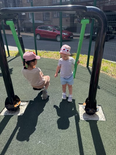 The image captures two young girls on a playground in the city of Vyazniki, Russia, on a sunny day. One girl, wearing a pink baseball cap and a beige t-shirt with beige pants, is seated on a swing and appears to be in motion. The other girl, sporting a pink hat adorned with hearts and a striped t-shirt with light blue shorts, stands beside the swing, holding one of the green support poles. Both girls are wearing light-colored sneakers. The playground features a rubberized green surface and is enclosed by a dark metal fence. In the background, a red car is parked on a street adjacent to a brick building. The shadows of the girls and the swing set are cast on the playground surface, indicating a bright sunlit environment.