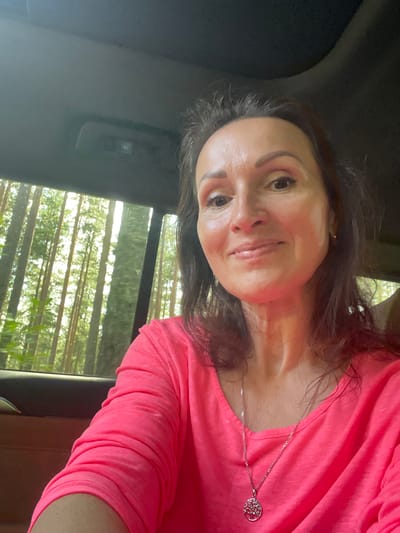 A woman is taking a selfie inside a car, with a forest visible through the window. The woman is in the foreground, wearing a bright pink long-sleeved shirt and a silver necklace with a tree pendant. She has dark hair and is smiling. The background shows a dense forest with tall trees, indicating a natural, outdoor environment. The image appears to be taken during daylight. The presence of a car interior suggests the woman is in transit or parked within the natural setting. There is no indication of the specific location within Solov'evka, Russia, other than the general description of a forest environment.