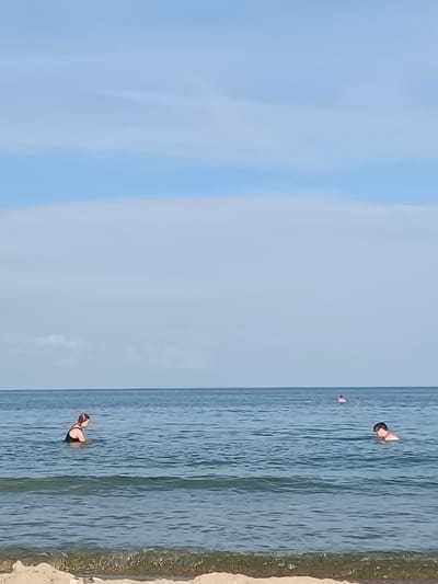 The image depicts a serene scene at the Black Sea coast in Zatoka, Ukraine. The foreground features a sandy beach with gentle waves lapping at the shore, indicating a calm and pleasant day. The water is a deep blue, transitioning to a lighter turquoise closer to the shore, with subtle ripples and small waves visible.
In the middle ground, two individuals are seen swimming in the sea. On the left, a person in a black swimsuit is partially submerged, facing left. On the right, another person, likely a male, is also in the water, wearing goggles. Further in the distance, two more figures are faintly visible, suggesting other beachgoers are enjoying the water.
The background is dominated by a vast expanse of the sky, which is a pale blue with soft, wispy white clouds scattered across it. The horizon line where the sea meets the sky is clear and distinct. The overall impression is one of leisure and relaxation on a sunny day at the beach.