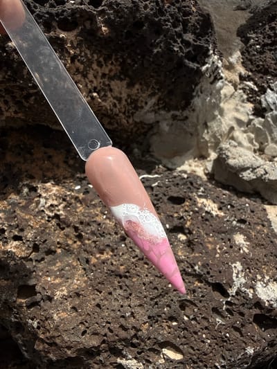 A nail sample is presented against a backdrop of volcanic rock, likely found in Yaiza, Spain. The nail is divided into three distinct sections: a light beige or nude color near the base, blending into a white section with glitter accents, and culminating in a light pink tip. The sample is attached to a clear, flat plastic holder. The volcanic rock behind the nail features a dark, porous texture typical of volcanic formations. The overall setting suggests a focus on showcasing the nail design against the backdrop of the island's unique geological features.