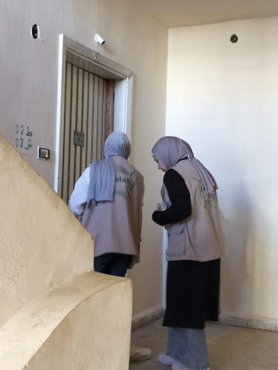 In an indoor setting within a building in قرية الضبية, Syria, two individuals, likely women, are captured standing on a light-colored floor, positioned near a closed door. Both individuals wear light-colored vests with a green logo/text, possibly "امان" (Aman, meaning 'safety' or 'security'), and headscarves (hijabs).
The individual on the left wears a light blue/grey hijab, a white long-sleeved shirt under the vest, and dark trousers; their back is mostly towards the viewer, facing a closed, vertically paneled metal door with a white frame. Above this door, a white pipe or conduit protrudes downwards, and to its left, a dark circular hole and an electrical switch/outlet are visible on the light-colored wall. Handwritten numbers "02" and Arabic characters "دل 02 ش" are marked on the wall near the door.
The individual on the right wears a light grey hijab, a black long-sleeved top under the vest, and a long black skirt over light blue jeans; they are angled slightly, looking downwards. A prominent, light-colored concrete stair railing occupies the foreground from the lower left. The walls are light-colored, and another dark circular hole is visible on the wall to the right. The scene depicts a functional indoor space, likely a stairwell landing or hallway.