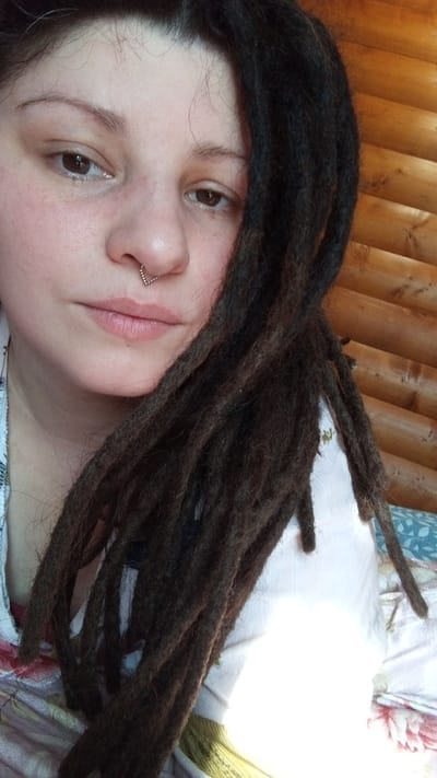 A close-up shot shows a person with dark brown dreadlocks partially covering their face. The person has light skin, brown eyes, and a septum piercing in their nose. They wear a patterned top in shades of white, pink, and green. Behind the person is a wood-paneled wall. The image appears to be taken in a well-lit indoor setting. It is not possible to confirm the location as Юдино, Russia, based solely on the visual content of the image.
