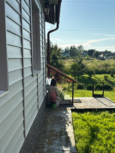 In the village of Bolshe Ozerki, Russia, a young child, facing away from the camera, stands on a wet, concrete walkway beside a light-colored, horizontal-slatted house. The child wears a patterned long-sleeved shirt, pink pants, and green boots, and is holding a white plush toy. A white pot with flowers is next to the child. The walkway leads to a raised wooden deck with a metal railing. Beyond the deck, a lush green lawn stretches towards a rural landscape with trees, bushes, and scattered houses under a clear blue sky. The scene appears to be a residential area on a sunny day.