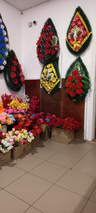 The image depicts a retail display of artificial flowers, primarily funeral wreaths and bouquets, likely for sale in Ertis, Kazakhstan. The setting appears to be an indoor establishment with white walls and a tiled floor.
Dominating the upper portion of the image are several funeral wreaths of various designs and colors, predominantly green, red, and black, with some featuring gold accents and religious symbols like a cross. These wreaths are affixed to the wall.
Below the wreaths, a large assortment of artificial flowers in vibrant colors—including red, pink, yellow, orange, and white—are arranged in cardboard boxes and possibly buckets. Some of these are stacked, suggesting a commercial arrangement for display and sale. One box in the lower right corner has Cyrillic writing and the number "450". A security camera is visible on the upper left wall.
The overall scene is a still-life presentation of merchandise in what appears to be a flower shop or a similar retail environment. There are no people visible in the image.