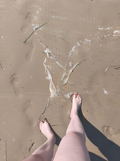 The image shows a person's bare feet standing on wet sand at the beach in San Salvo, Italy. The sand is a light brown color and appears damp, with some footprints visible. A small amount of white sea foam and some green seaweed are also present on the sand. The person's toenails are painted pink. The sun is casting a shadow of the person's leg onto the sand.