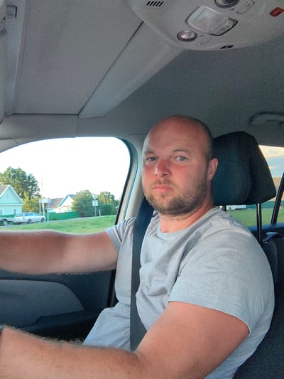 The image shows a man with a bald head and a beard, wearing a light gray t-shirt, sitting in the driver's seat of a car. He is looking directly at the camera. The interior of the car is visible, including the ceiling with lights and a sunroof. Outside the car window, a rural landscape with houses and trees can be seen. The man is wearing a black seatbelt. The scene appears to be captured during daytime, possibly late afternoon due to the lighting. The location is identified as Dzyarzhynsk, Belarus.
