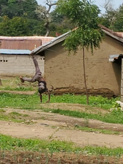 The image shows a young person in the process of a cartwheel or similar acrobatic move. They are wearing a dark t-shirt with a graphic design and light-colored pants. The person is mid-air, with one leg extended upwards and the other bent, and their arms are outstretched to the sides. They appear to be on a dirt path or ground next to a building. The building has a textured, earthy-colored wall and a slanted roof, with some open doorways or windows visible. Lush green vegetation, including trees and grass, surrounds the area, suggesting an outdoor setting in a natural or semi-rural environment. The lighting suggests it is daytime, possibly late morning or afternoon, and the weather appears clear. The overall scene is informal and suggests an act of play or athletic demonstration.