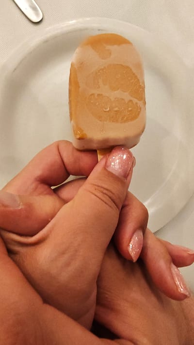 A hand with light pink, glittery nail polish holds a popsicle that has a lipstick imprint on it. The popsicle is light brown and appears to be melting, with drips of liquid visible. The hand is positioned over another hand, also with a similar nail polish color. They are resting on a white tablecloth, with a white plate and a sliver of a metal utensil visible in the background. The setting appears to be indoors, possibly a restaurant or dining area, given the tablecloth and plate. The lighting suggests it could be daytime. The overall impression is one of leisure and indulgence. The lipstick imprint on the popsicle adds a playful and intimate touch. No visible text. The location context is Litohoro, Greece.