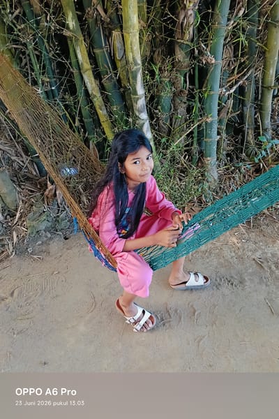A young girl with long, dark hair is sitting in a hammock made of rope and netting. She is wearing a pink outfit consisting of a long-sleeved shirt and pants. She has white sandals on her feet. The hammock is suspended between bamboo stalks, which are visible in the background. The ground beneath the hammock appears to be dirt. The scene is lit by natural sunlight. The image includes a watermark at the bottom left corner that reads "OPPO A6 Pro 23 Juni 2026 pukul 13.03". This suggests the photo was taken on June 23, 2026, at 1:03 PM, and was captured with an OPPO A6 Pro phone. The mention of "Rheum Barat, Indonesia" provides a specific geographical location for the scene.