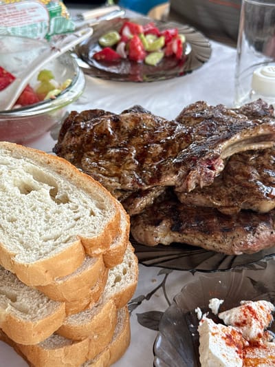This is an eye-level, close-up, outdoor, still life photograph taken in Ravadinovo, Bulgaria, depicting a meal spread on a white tablecloth. The foreground is dominated by a stack of several thick slices of white bread, with a crusty, golden-brown exterior and a soft, airy interior. To the right of the bread, a pile of grilled steaks, appearing to be ribeye, is presented on a dark, translucent plate. The steaks are well-seared, with visible grill marks and a glistening, juicy appearance. In the background, to the left, a glass bowl contains a chopped salad of tomatoes and cucumbers. Further to the right, another dark, translucent plate holds chunks of white cheese, possibly feta, generously sprinkled with paprika. A clear glass and a white salt shaker are also visible in the background. The overall scene suggests a casual outdoor dining experience.
