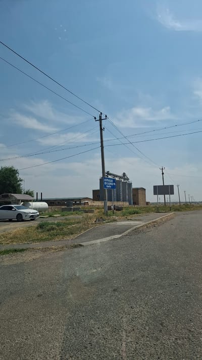 A street scene in the city of Қасымбек датқа, Kazakhstan, is captured under a bright, clear sky with scattered clouds. The foreground features an asphalt road leading towards the right side of the frame, with a curb and a strip of dry grass on the left. A white sedan is parked on the left side of the road. In the middle ground, a blue directional sign is mounted on a pole, indicating "МОЛЫБАЙ ОРАЗАЛИЕВ → АУЫЛЫ" which translates to "MOLYBAI ORAZALIEV → VILLAGE." Behind the sign, a large industrial complex with several tall, silver silos and a brick building is visible. Power lines stretch across the sky, supported by utility poles. The overall environment appears to be semi-rural or industrial.