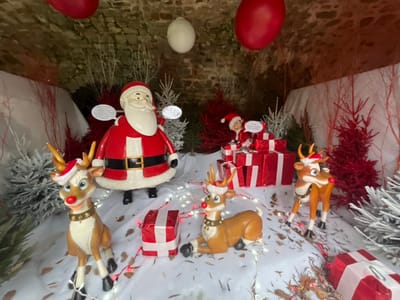An indoor Christmas display is arranged within a stone-arched alcove in Bagnoles de l'Orne Normandie, France. The ground is covered with artificial white snow, illuminated by scattered red string lights and interspersed with brown leaf debris. White fabric drapes form a snowdrift-like backdrop.
Central to the scene is a large Santa Claus figure, standing, dressed in a red suit with white trim, a black belt with a gold buckle, and black boots. A white speech bubble is positioned near his head. To the right, a smaller elf figure in a red suit and hat perches atop a stack of red gift boxes with white ribbons, also featuring a white speech bubble.
Four reindeer figures populate the display: one lies in the left foreground, adorned with a red nose, gold antlers, a red Santa hat, and a gold bell collar. A second stands to its right, similarly equipped with a red nose, gold antlers, and a red Santa hat. A third stands near the center-right, showing a red nose and gold antlers. A fourth stands on the far right, distinguished by a red nose, antlers, and a black harness.
Multiple artificial Christmas trees are arranged throughout, some frosted white and others solid red. Additional bare red branches are integrated into the scenery. Several individual red gift boxes with white ribbons are placed on the ground. Three spherical ornaments hang from the stone ceiling: two red and one white.