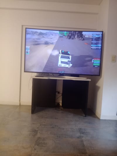 The image depicts a room setting with a television mounted on a white wall. The TV is displaying a video game, showing a vehicle moving along a terrain. A black and white TV stand is present below the TV. The floor is covered with light grey tiles. The room appears to be illuminated by artificial light. The location, Lomas de Zamora, Argentina, is not discernible from the visual information. There are no people visible in the image, nor any interactions taking place within the frame.