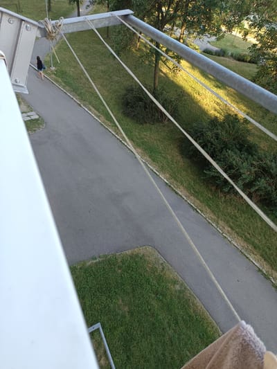 An elevated, high-angle, medium shot captures a view of a paved pathway with a person walking on it, bordered by grassy areas and trees. The scene appears to be outdoors, possibly in a park or residential area. A metal structure with wires runs diagonally across the frame, from the upper left to the lower right. The person, seen from behind and walking away from the camera, is a woman wearing a dark top and light-colored shorts, carrying a blue bag. The pathway is asphalt, and the surrounding ground is covered in grass, with some areas appearing dry. Sunlight casts shadows from the trees and the metal structure onto the ground. A beige towel hangs into the frame from the bottom right corner. The image location is identified as Košice, Slovakia.