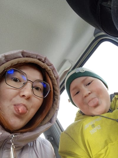 The image displays two individuals, an adult and a child, inside what appears to be the cabin of a vehicle. Both subjects are captured in a close-up, selfie-style photograph, facing the camera with their tongues extended.
The individual on the left, an adult female, has reddish-brown hair partially covered by a light purple hooded jacket with a beige inner lining. She wears eyeglasses with dark frames, reflecting blue light sources. A silver zipper pull is visible on her jacket. The individual on the right, a child, wears a green and white striped knit beanie and a bright yellow hooded jacket. Text, possibly "SPORT" and a circular logo, is visible on the child's jacket, appearing reversed.
The background shows the light grey/beige interior ceiling of the vehicle, dark interior trim on the upper right, and a window revealing an overcast, bright sky. The specific location within the city of Старый Шагирт, Russia, is not discernible from the visual information provided within the image.