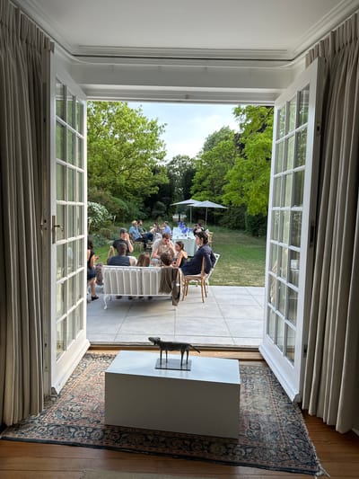 An open double French door, flanked by beige curtains, frames a view of an outdoor gathering. The door opens onto a tiled patio, transitioning to a grassy lawn with scattered trees and flowering bushes. A group of people are seated at tables set up with white cloths and blue flowers, under a white parasol. Inside, a rectangular white plinth stands on a patterned rug near the door; atop it is a bronze statue of a quadruped, possibly a deer or antelope. The room has wooden flooring. This scene is likely in Cambridge, United Kingdom, due to the architecture style and landscape.