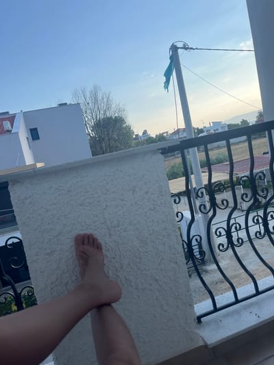 The image shows a view from a balcony in Ofrynio Beach, Greece. In the foreground, a person's legs and feet are visible, resting on a white textured surface. The person appears to be barefoot. Beyond the balcony railing, a street or yard area is visible, with buildings and trees in the background. A utility pole with a blue flag attached to it stands in the middle ground. The sky is clear with some scattered clouds.