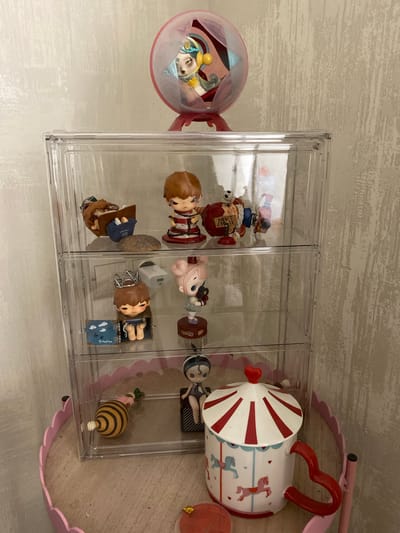 In Lymanka, Ukraine, a clear plastic three-tiered display case sits on a pink scalloped decorative shelf. The case contains several small figurines. On the top shelf, a figure resembling a child with brown hair lies on its side reading, and another figure with brown hair and a red and white striped outfit is bound with rope. A third figure, a clown-like character with a sign that reads "DON'T CROSS," is also on this shelf. The middle shelf holds a figure with a crown and a small open box with clouds and the word "Freedom" written on it, as well as a figure with pink hair. The bottom shelf contains a bumblebee-shaped object and a figurine dressed in black and white. To the right of the display case, a ceramic mug with a red and white striped lid, decorated with carousel horses, is prominently featured. Above the display case, a pink spherical container holds a single figurine. The background is a textured light-colored wall.