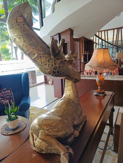 A brass giraffe sculpture with a smaller giraffe head nestled near its face rests on a wooden table. The giraffe's long neck arches upwards. A blue velvet chair and a small potted plant sit on a nearby table. In the background, there is a lamp with a fringed shade, a staircase with wood banisters, and a view of trees through a window. The scene appears to be a hotel lobby or a well-decorated living room.