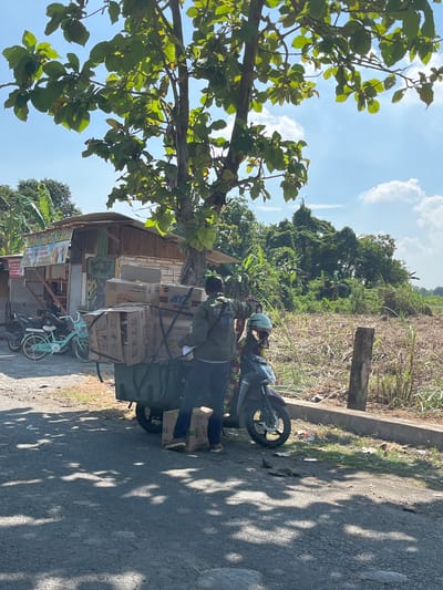 In Pamotan, Indonesia, a man in a green jacket and dark pants stands next to a motorcycle with a trailer filled with boxes. The trailer is attached to the motorcycle's rear wheel. The motorcycle also carries a helmet. The man appears to be in the process of handling the boxes. In the background, there is a small wooden building with a corrugated metal roof, possibly a shop, and a tree with large green leaves. Beyond the building and the road, there is a field with dry vegetation and a dense area of green trees. The sky is blue with scattered white clouds. A light blue bicycle is parked to the left of the trailer. The scene is lit by sunlight, casting shadows on the asphalt road.